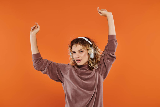 mujer rizada encantadora y feliz en auriculares inalámbricos escuchando música y bailando en naranja - Foto, imagen