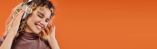 pleased curly woman in wireless headphones listening music with closed eyes on orange, banner - Photo, Image