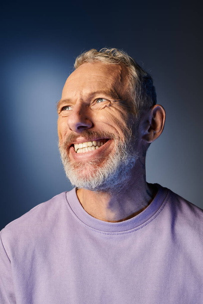 wesoły brodaty dojrzały mężczyzna w luźnej purpurowej bluzie z akcesoriami uśmiecha się i odwraca wzrok - Zdjęcie, obraz