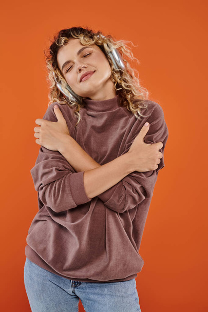 dreamy woman with wavy hair listening music in wireless headphones and hugging herself on orange - Photo, Image