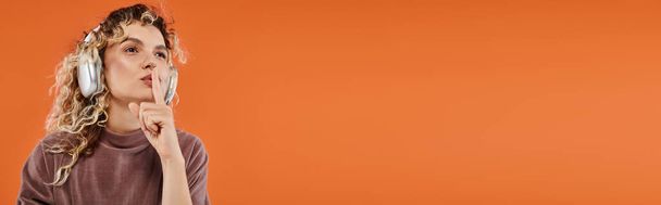 stylische lockige Frau, die Musik in drahtlosen Kopfhörern hört und Schweigezeichen auf orangefarbenem Banner zeigt - Foto, Bild