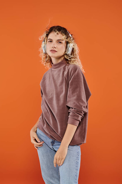 expressive woman with wavy hair in brown turtleneck listening music in wireless headphones on orange - Photo, Image