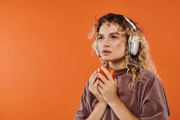 mujer rizada pensativa con taza de café de la mañana escuchando música en auriculares inalámbricos en naranja - Foto, Imagen