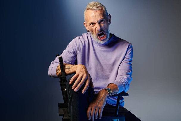 stylish mature man in purple sweatshirt sitting on chair while grimacing and looking at camera - Photo, Image