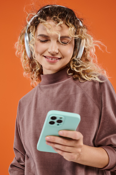 glücklich gelockte Frau schaut auf Smartphone und hört Musik in drahtlosen Kopfhörern auf orange - Foto, Bild