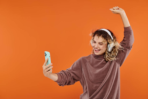 ワイヤレスヘッドフォンでダンスし,オレンジ色でスマートフォンでセルフィーを取る陽気なカーリー女性 - 写真・画像