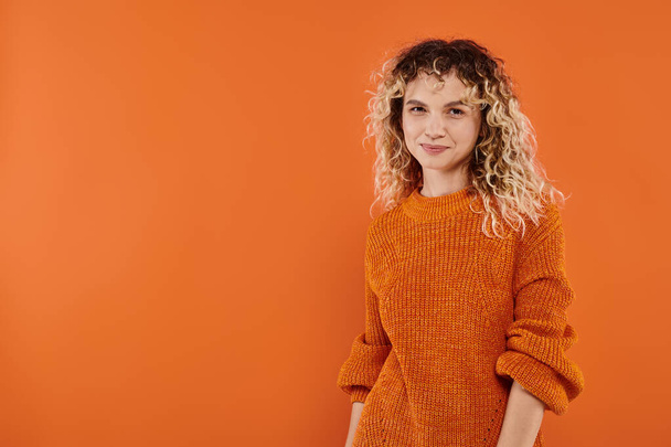 stylish woman with wavy hair in cozy knitted sweater looking at camera on bright orange backdrop - Foto, immagini