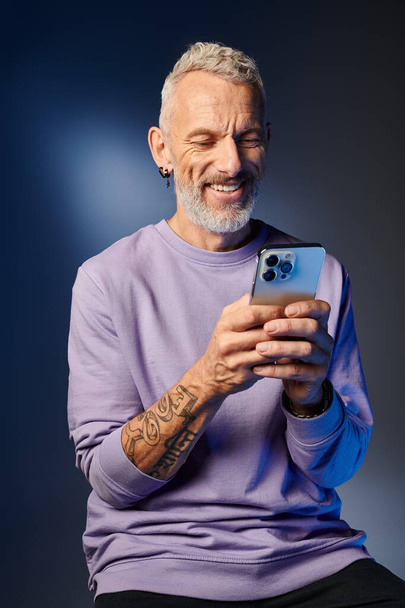 vidám jóképű, érett férfi lila pulóverben, szürke szakállal nézi az okostelefonját. - Fotó, kép