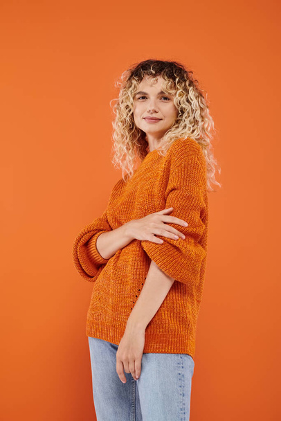 trendy woman with wavy hair in cozy knitted sweater looking at camera on bright orange backdrop - Фото, изображение