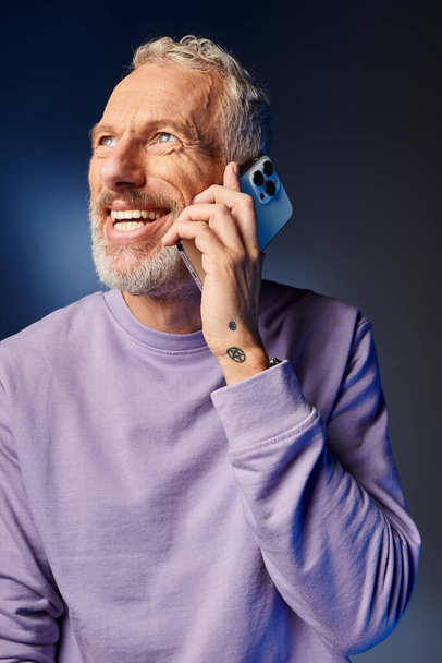 alegre barbudo hombre maduro de moda en sudadera elegante púrpura hablando por teléfono sobre fondo azul - Foto, imagen