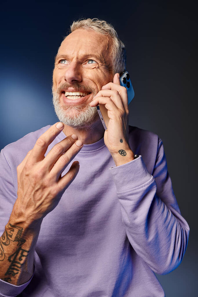 vrolijk baard trendy volwassen man in paars stijlvol sweater praten per telefoon op blauwe achtergrond - Foto, afbeelding