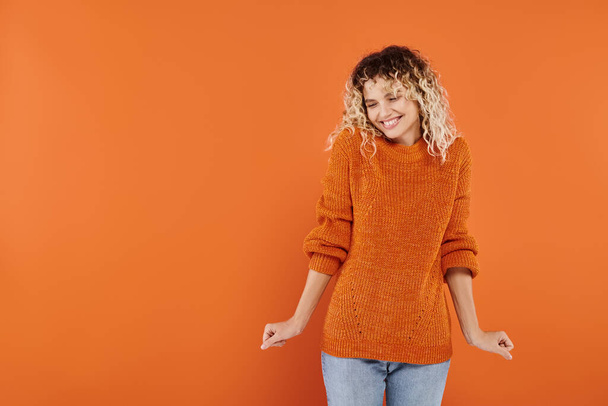 vidám és göndör nő fényes kötött pulóverben mosolyog és pózol narancssárga stúdió háttérben - Fotó, kép
