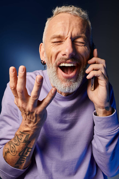 весёлый бородатый зрелый мужчина в фиолетовой стильной толстовке разговаривает по телефону на синем фоне - Фото, изображение
