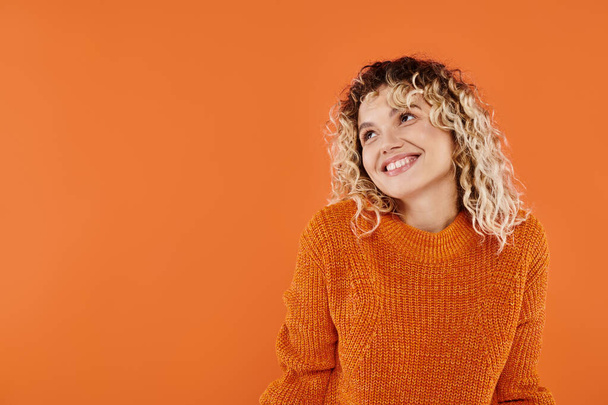ευτυχισμένη και σγουρή γυναίκα σε φωτεινό πλεκτό πουλόβερ χαμογελώντας κοιτάζοντας μακριά σε πορτοκαλί φόντο στούντιο - Φωτογραφία, εικόνα
