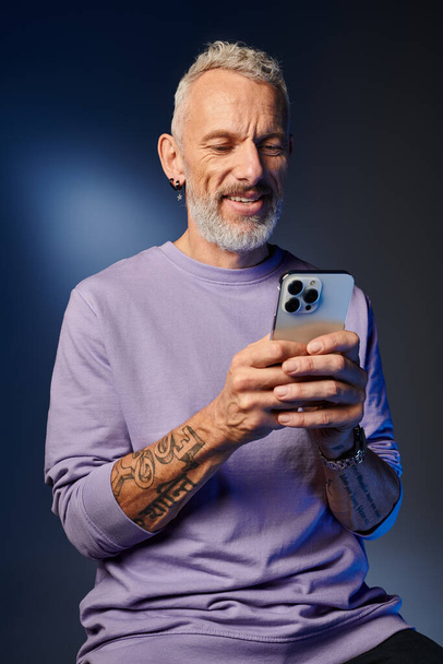χαρούμενος όμορφος ώριμος άντρας με μωβ μπλούζα και γκρι γενειάδα κοιτάζει το smartphone του - Φωτογραφία, εικόνα