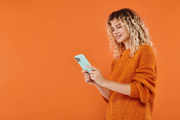 Lächeln lockige Frau in hellem Strickpullover surft im Internet auf Smartphone auf orangefarbenem Hintergrund - Foto, Bild