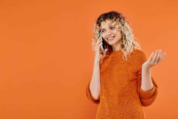明るいオレンジ色の背景で携帯電話で話す暖かい編まれたセーターで幸せなカーリー女性 - 写真・画像