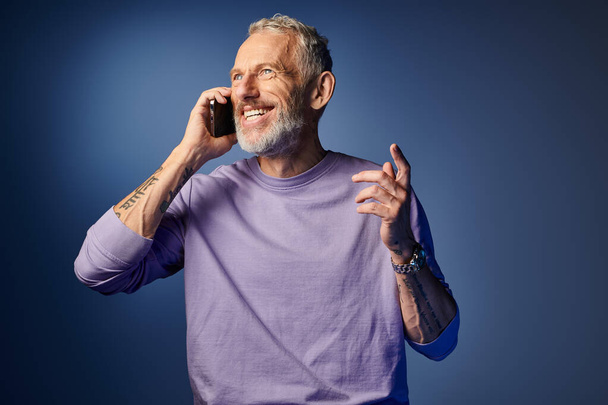 alegre barbudo hombre maduro de moda en sudadera elegante púrpura hablando por teléfono sobre fondo azul - Foto, imagen