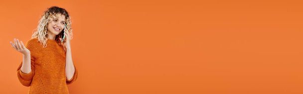 iloinen kihara nainen viihtyisässä neulottu villapaita puhuu matkapuhelimella kirkkaan oranssilla taustalla, banneri - Valokuva, kuva