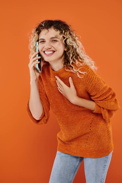 nadšené kudrnaté ženy v pleteném svetru mluví na mobilním telefonu a směje se na oranžovém pozadí - Fotografie, Obrázek