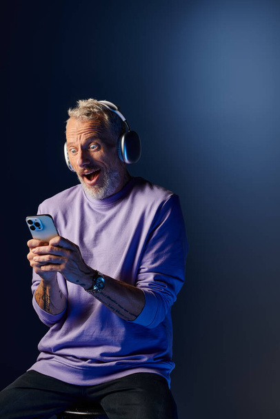 joyful surprised mature man with beard and headphones enjoying music and looking at his phone - Foto, immagini