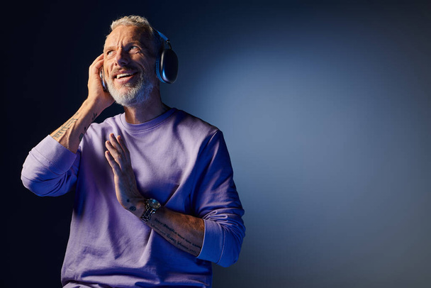 radosny atrakcyjny dojrzały mężczyzna z szarą brodą i słuchawkami w fioletowej bluzie cieszący się muzyką - Zdjęcie, obraz