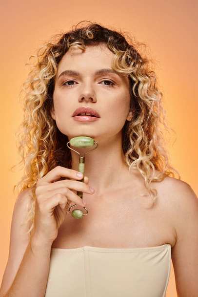 sensual mujer rizada con piel radiante usando rodillo de jade y mirando a la cámara en el fondo pastel - Foto, Imagen