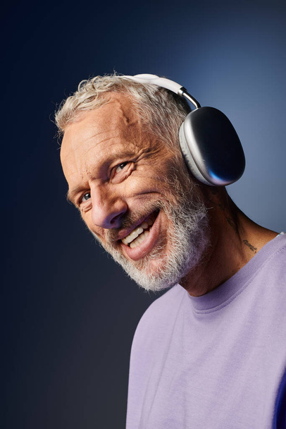 vrolijk knap volwassen man in paars sweatshirt met hoofdtelefoon genieten van muziek en kijken naar camera - Foto, afbeelding