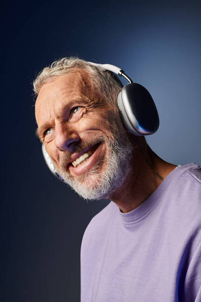 wesoły przystojny Dojrzały mężczyzna z szarą brodą i słuchawkami w fioletowej bluzie cieszący się muzyką - Zdjęcie, obraz