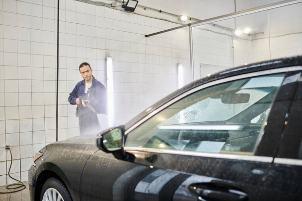 atrakcyjny dedykowany profesjonalista w niebieskim mundurze używający węża do mycia nowoczesnego czarnego samochodu w garażu - Zdjęcie, obraz