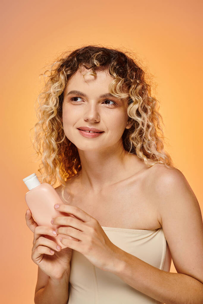 Kıvırcık saçlı neşeli kadın pastel arkaplanda vücut losyonuyla şişe tutuyor. - Fotoğraf, Görsel