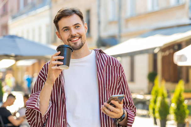 Szczęśliwa brunetka brodatego mężczyzny cieszącego się poranną kawą gorącym napojem i uśmiechem na świeżym powietrzu. Relaksuję się. Facet spacerujący ulicą miejskiego słońca, pijący kawę na wynos. Miejski styl życia na zewnątrz - Zdjęcie, obraz