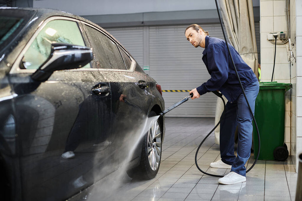 handsome hard working professional in blue comfy uniform with collected hair washing black car - Photo, Image