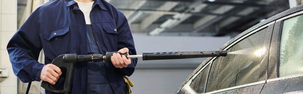 περικοπή άποψη του σκληρά εργαζόμενου επαγγελματία σε μπλε στολή χρησιμοποιώντας σωλήνα για να πλύνετε μαύρο αυτοκίνητο, πανό - Φωτογραφία, εικόνα