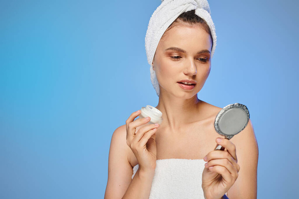 mulher encantadora com creme facial e toalha na cabeça olhando para o espelho no fundo azul, auto-cuidado - Foto, Imagem