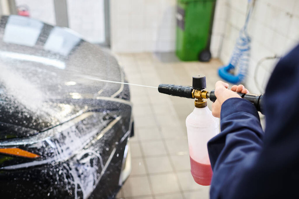καλλιεργημένη άποψη του αφοσιωμένου επαγγελματία με μπλε στολή χρησιμοποιώντας σαπούνι για να πλύνετε μαύρο σύγχρονο αυτοκίνητο - Φωτογραφία, εικόνα