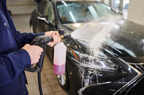 vista recortada de especialista dedicado al trabajo duro en uniforme azul usando jabón para lavar el coche negro - Foto, imagen