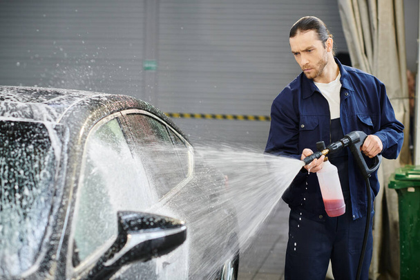 atrakcyjny skoncentrowany przystojny mężczyzna z zebranymi włosami w niebieskim mundurze mycie czarny samochód z mydłem - Zdjęcie, obraz