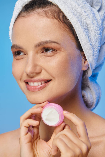 radosna kobieta ze świecącą skórą i ręcznikiem trzymająca słoik kosmetycznego kremu i odwracając wzrok na niebiesko - Zdjęcie, obraz