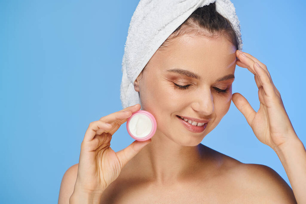 pleased charming woman with closed eyes and towel holding face cream and smiling on blue backdrop - Photo, Image