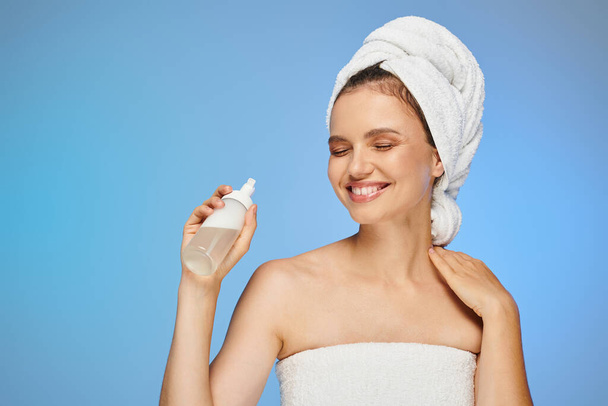 cheerful woman with towel on head and perfect face holding dispenser with cosmetic foam on blue - Photo, Image