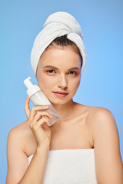mujer atractiva con toalla en la cabeza dispensador de sujeción con espuma facial mirando a la cámara en azul - Foto, Imagen