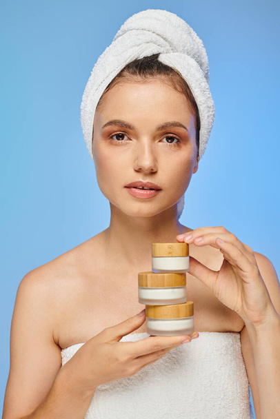 mulher atraente com pele limpa e toalha na cabeça segurando creme cosmético olhando para a câmera em azul - Foto, Imagem