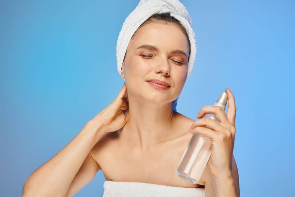 smiling woman with radiant skin and closed eyes holding bottle of body spray on blue backdrop - Photo, Image