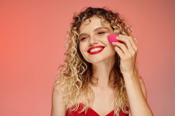 curly cheerful woman with red lips using face sponge on pink and yellow backdrop, elegant beauty - Photo, Image