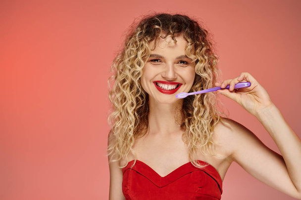 fröhliche lockige Frau mit roten Lippen und Zahnbürste, die vor pastellfarbenem Hintergrund in die Kamera blickt - Foto, Bild