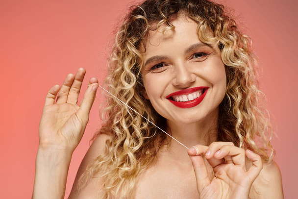 ritratto di donna felice con capelli ondulati e labbra rosse pulizia dei denti con filo interdentale su rosa - Foto, immagini
