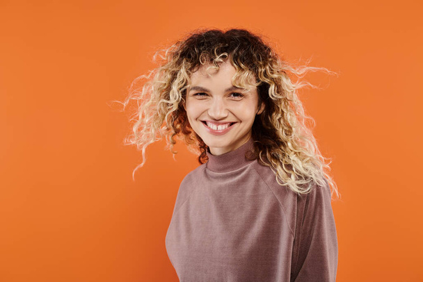 cheerful woman with wavy hair in mocha color turtleneck looking at camera on radiant orange backdrop - Photo, Image