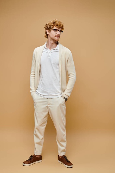 young redhead man in eyeglasses and old money style casual attire with hands in pocket on beige - Photo, Image