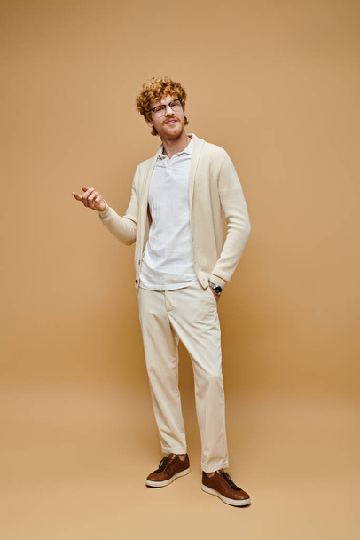 full length of elegant redhead man in eyeglasses and trendy clothes with hand in pocket on beige - Photo, Image
